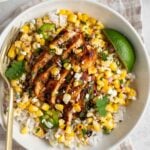 Close up of street corn chicken rice bowls with fresh cilantro and feta on top with a lime wedge on top with a gold fork of a gingham beige tea towel on a white background.