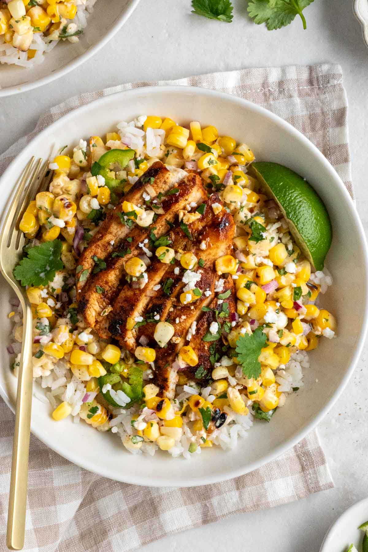 Close up of street corn chicken rice bowls with fresh cilantro and feta on top with a lime wedge on top with a gold fork of a gingham beige tea towel on a white background.