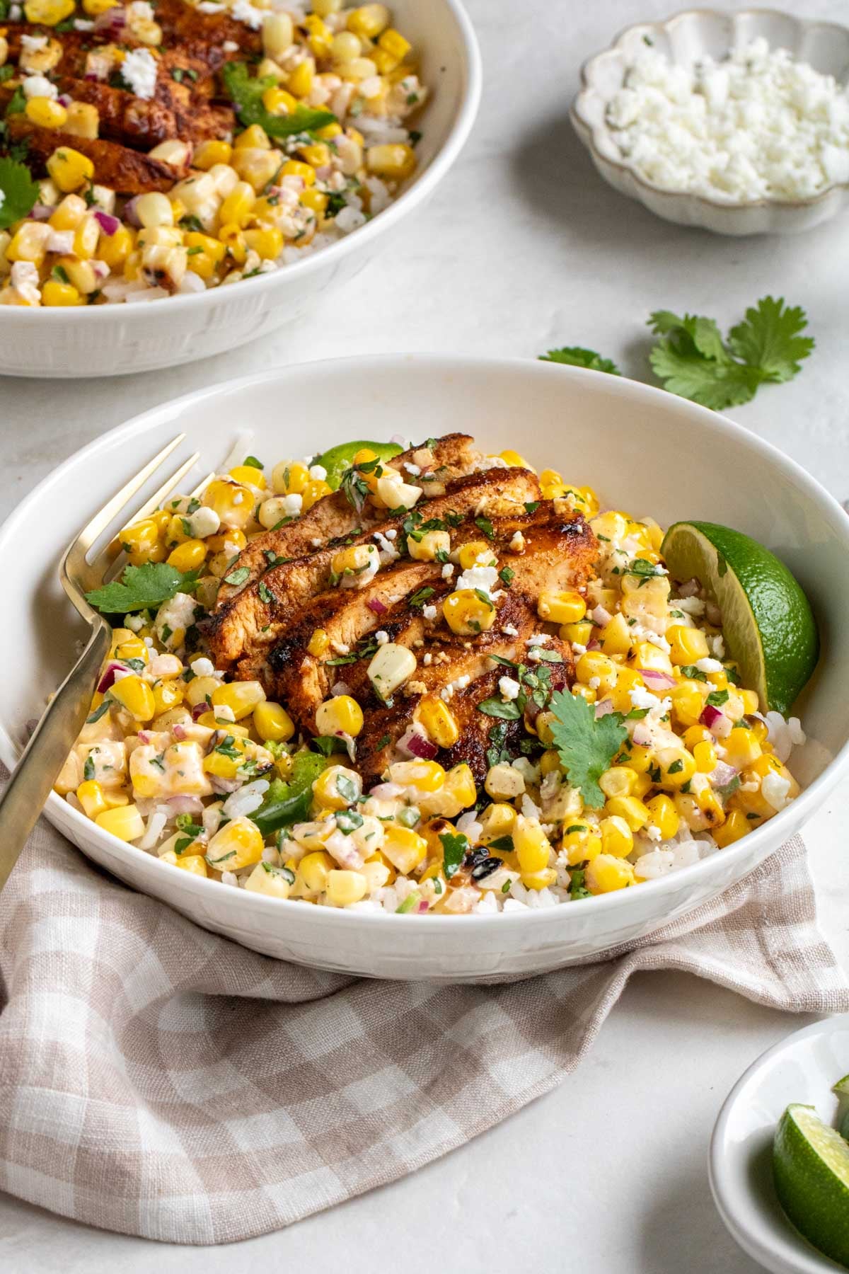 Close up two Mexican street corn chicken rice bowls on a white background with fresh cilantro and crumbed cheese in the background.
