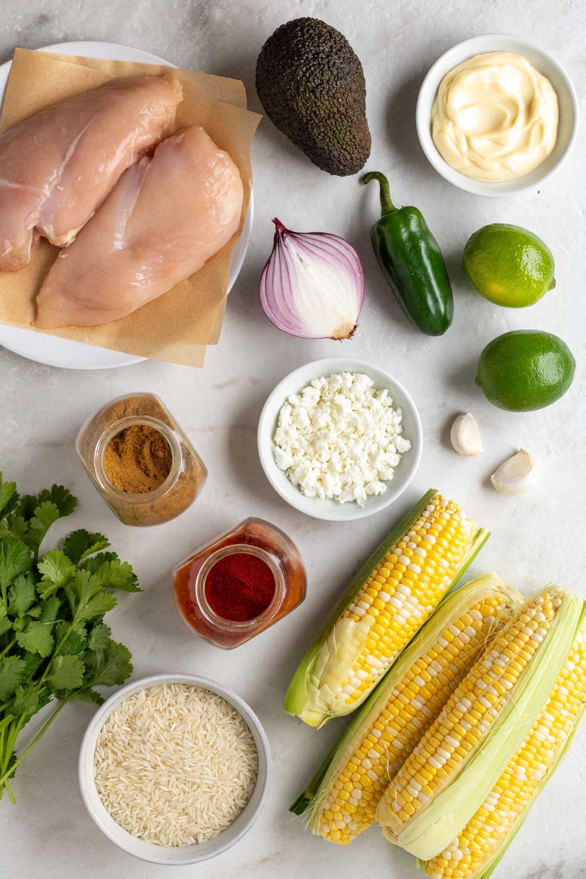 Chicken breasts on a white plate with small bowls of mayonnaise and crumbled cheese with avocado, red onion, jalapeno, limes, garlic cloves, cumin, paprika, corn ears, rice, and fresh cilantro on a white background.