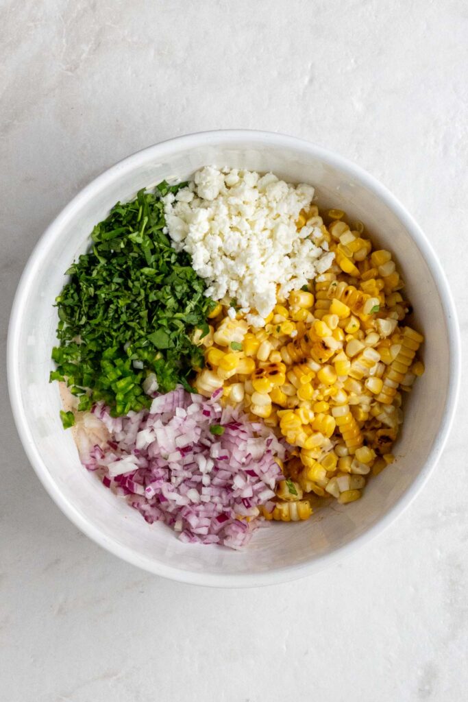 White bowls with grilled corn kernels, diced red onion, diced jalapeno, chopped cilantro, and crumbled cheese on a white background.