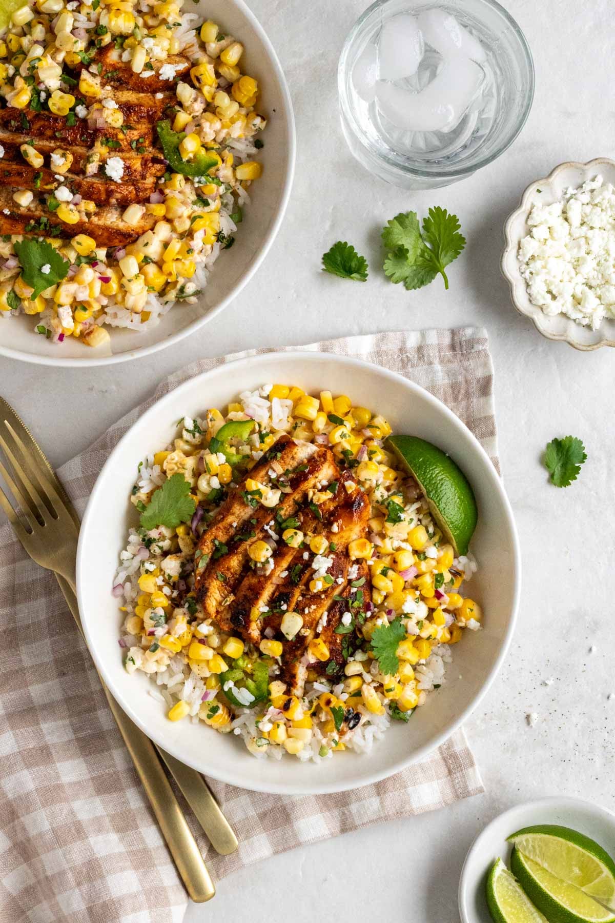 Two bowls filled with cooked rice, street corn salad, and grilled chicken with crumbled cheese, cilantro, and lime wedgest on the side on a tea towel with gold utensils and a glass of sparkling water on a white background.