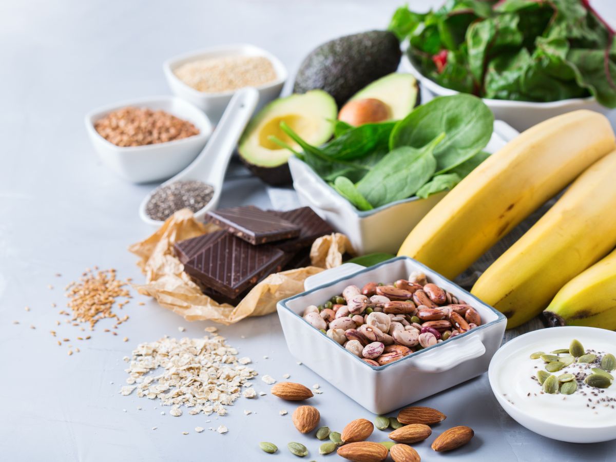 Bowl of yogurt with pumpkin seeds and chia seeds, bowl of beans and almonds, bowl of spinach, bowl of swiss chard, bowl oats, bowl of flax seeds, banana, dark chocolate, and avocados on a blue background.