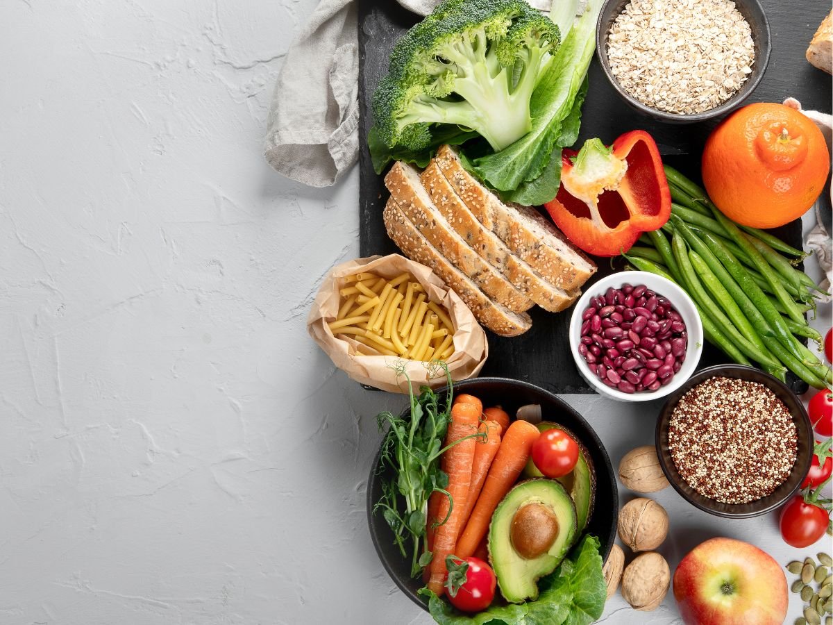 Display of broccoli, oats, lettuce, orange, bell pepper, whole wheat bread, brown rice pasta, kidney beans, green beans, quinoa, cherry tomatoes, sunflower seeds, apple, walnuts, avocado, and carrots on a dark cutting board on a light grey background with a grey tea towel.