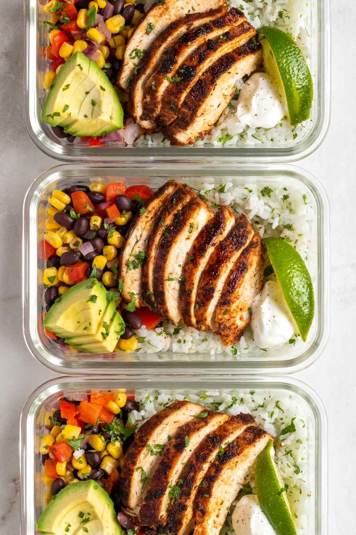 Close up of three Southwest chicken bowls in a glass meal prep container with sliced avocado, lime wedges, and chopped cilantro on top.