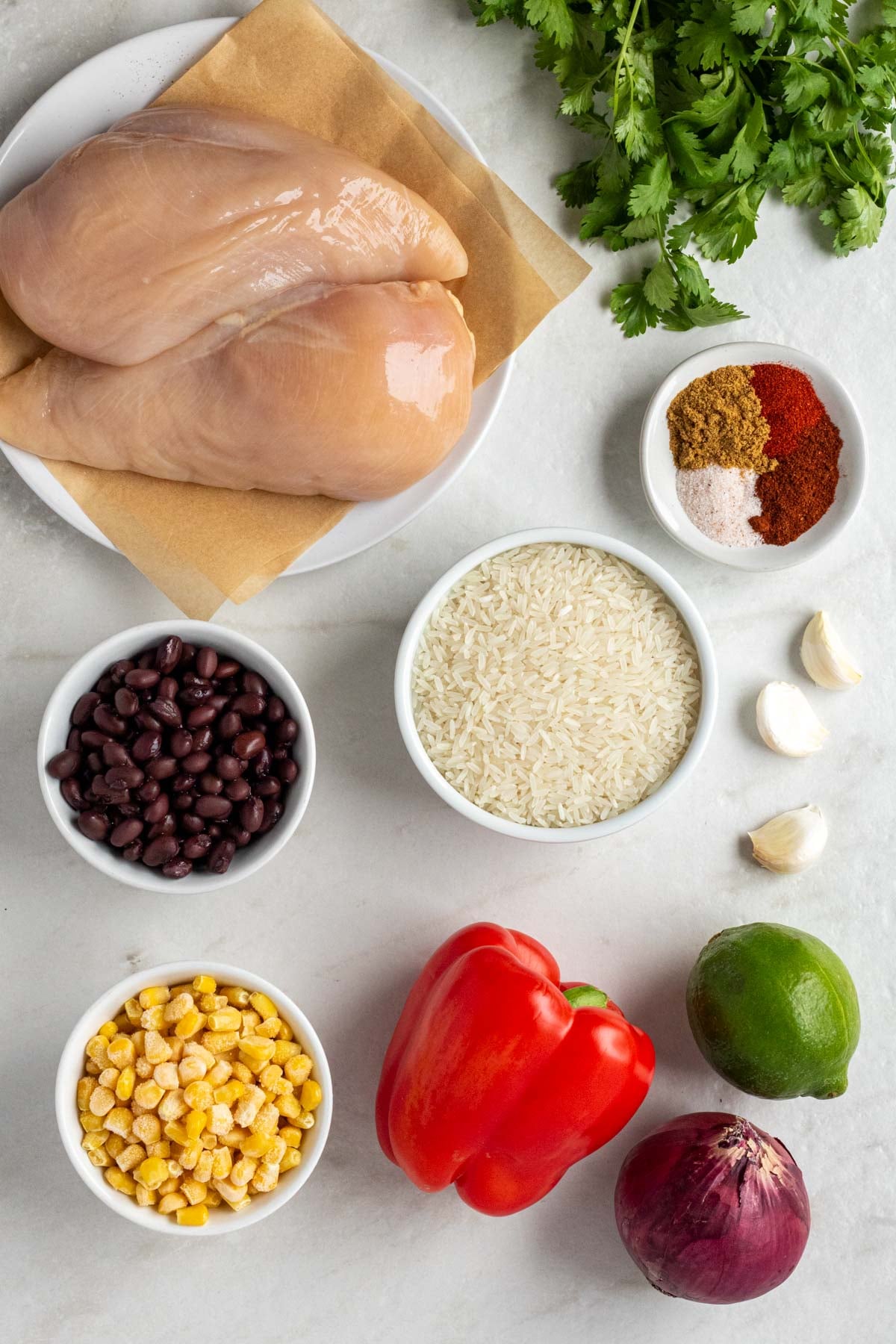 Two raw chicken breasts on a white plate with a bowl of white rice, black beans, frozen corn, Southwest spices, garlic cloves, red onion, red bell pepper, lime, and fresh cilantro. 
