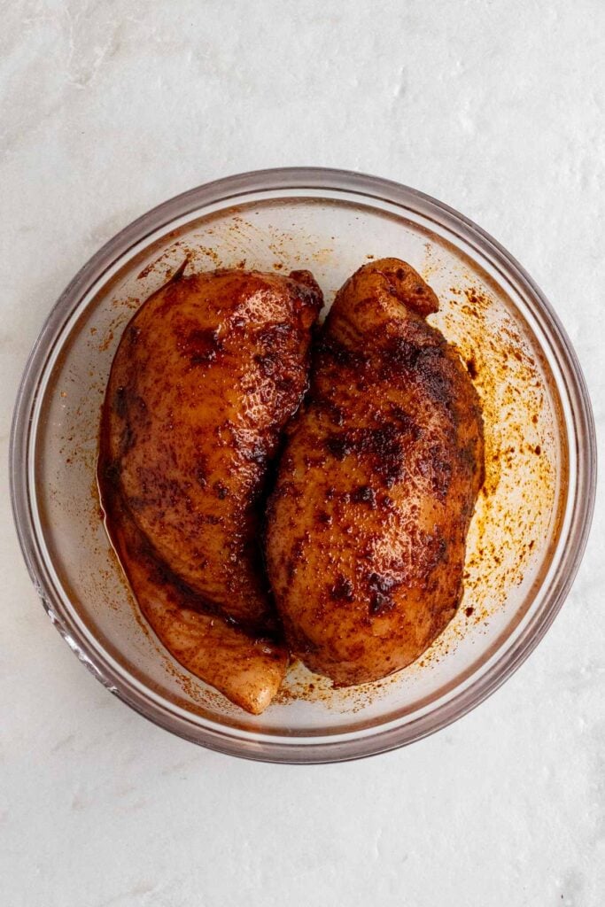 Two chicken breasts marinating in Southwest spices in a glass bowl.