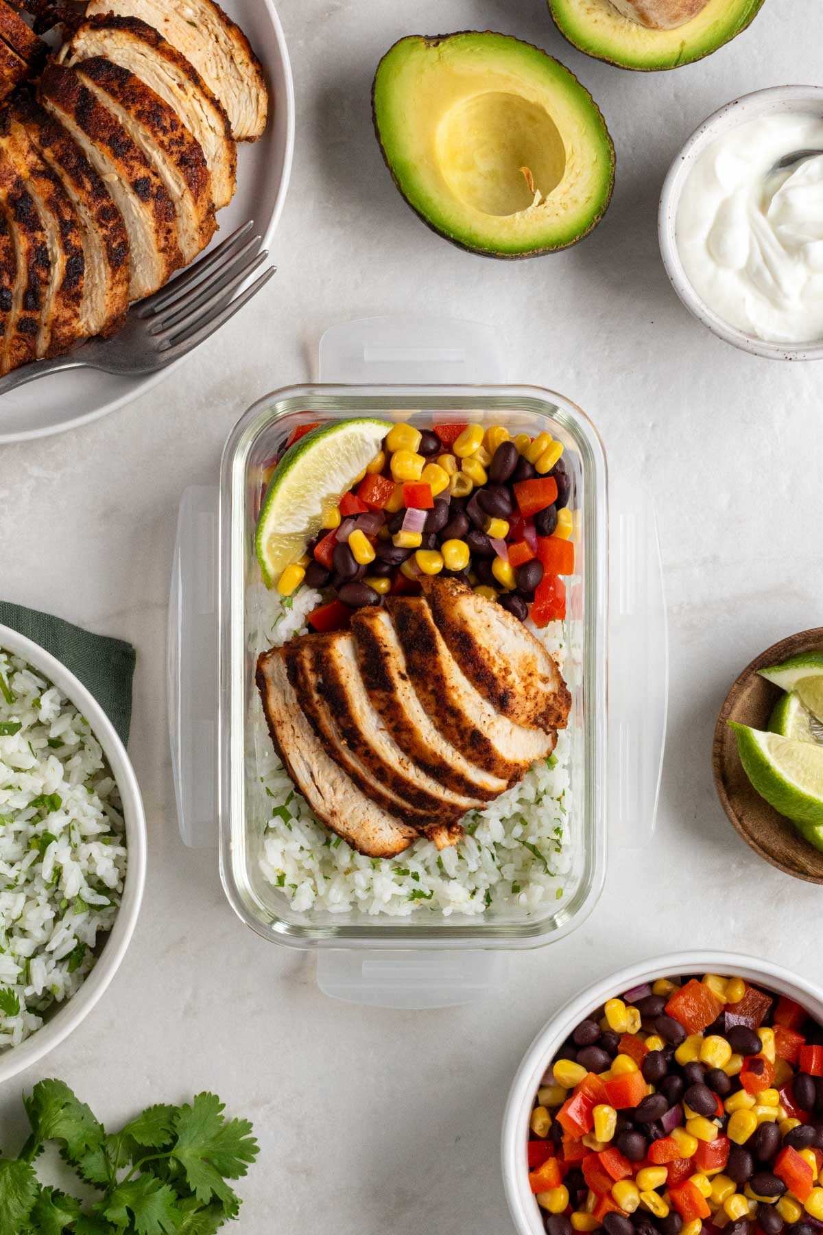 Glass meal prep container with rice, chicken, veggies with a sliced Southwest chicken, avocado, sour cream, cilantro lime rice, lime wedges, and a corn, black bean, and red pepper salad on the side on a white background.