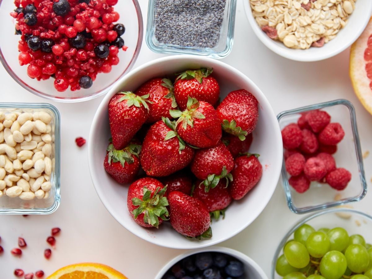 Big bowl of fresh strawberries, with smaller bowls of frozen berries, poppy seeds, rolled oats, raspberries, grapes, blueberries, peanuts, with half an orange and grapefruit on the side on a light pink background.