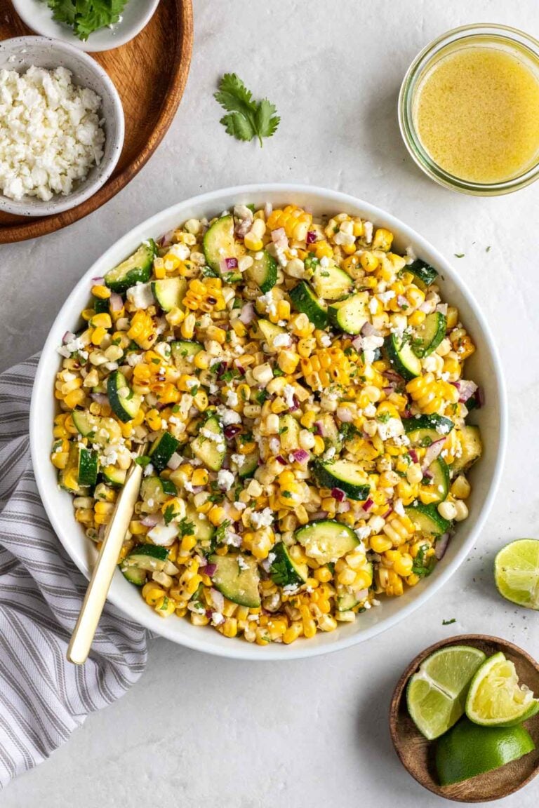 White bowl of grilled corn and zucchini salad with a gold serving spoon with a jar of salad dressing, and a bowl of crumbled feta, fresh cilantro, and lime wedges with a tea towel on a white background.