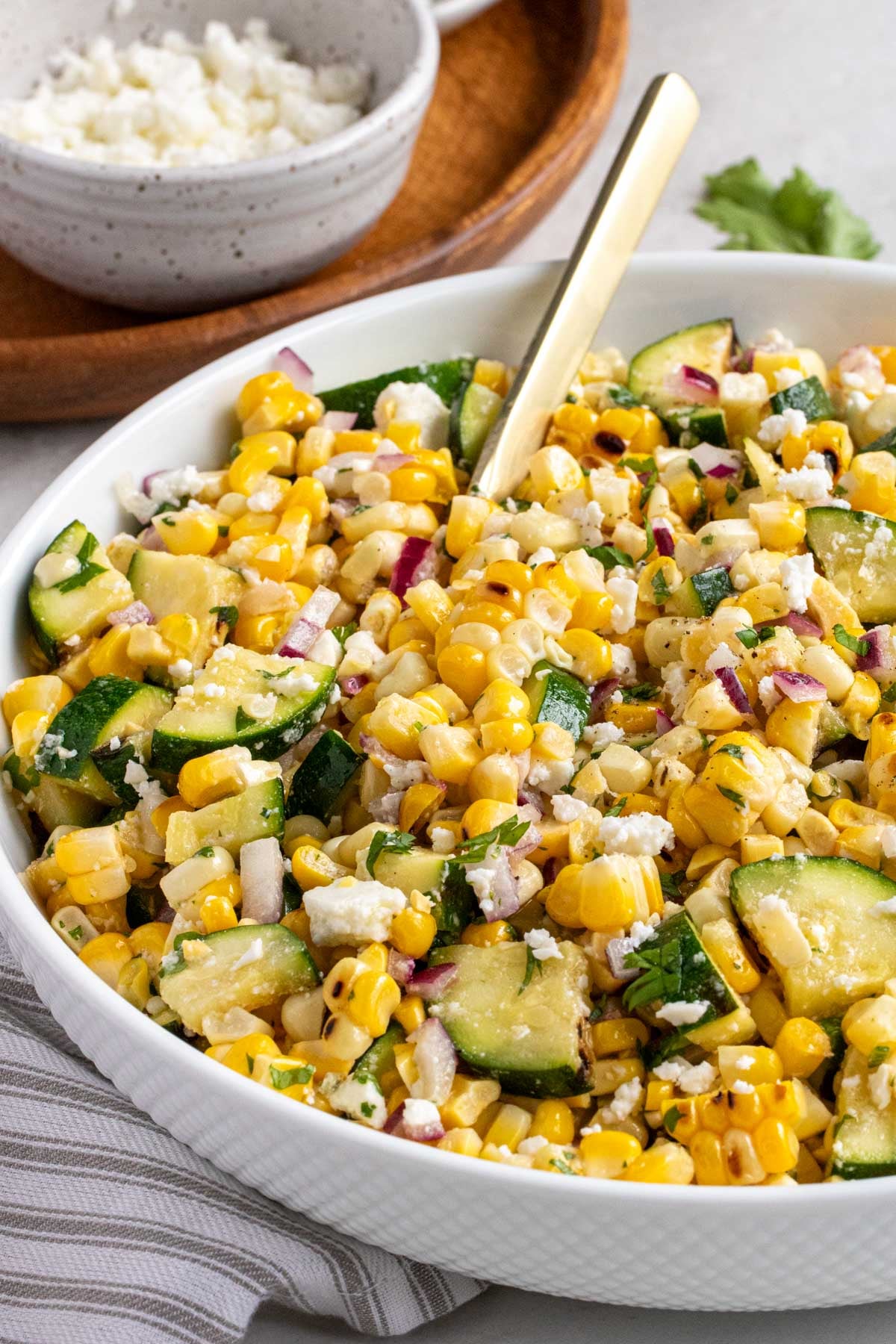 Close up of a bowl of corn and zucchini salad with grey and white striped tea towel and a bowl of feta cheese in the background.