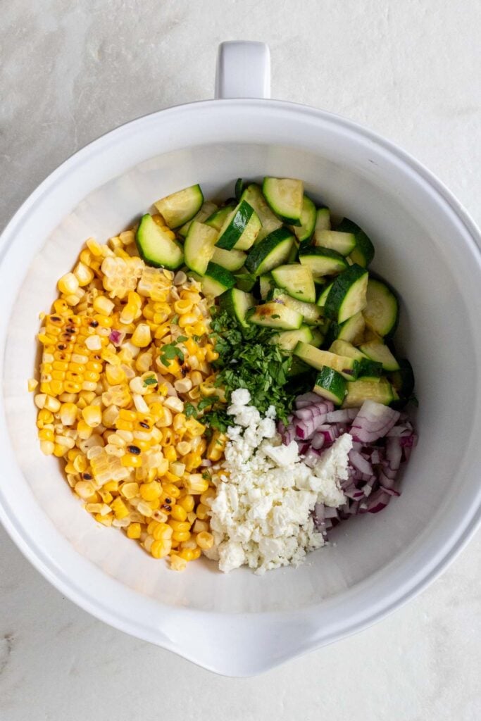 Large white mixing bowl with grilled corn kernels, diced grilled zucchini, diced red onion, crumbled feta, and chopped cilantro.