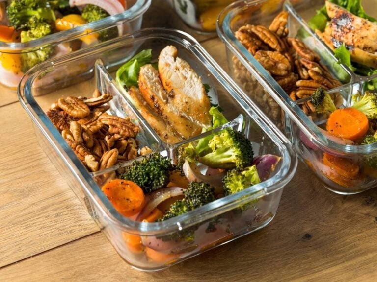Glass meal prep containers filled with cooked chicken thighs, roasted vegetables, and pecans on a wood table.