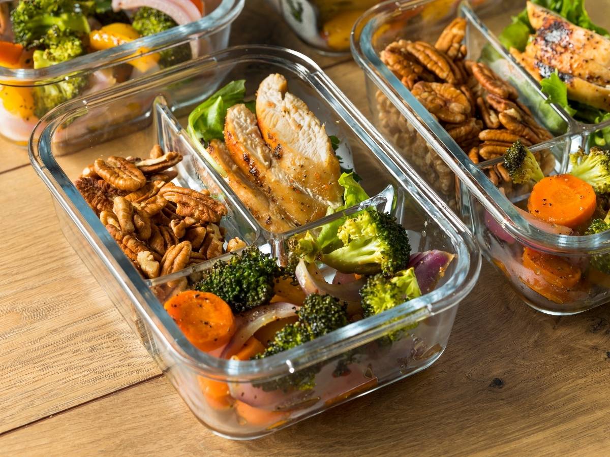 Glass meal prep containers filled with cooked chicken thighs, roasted vegetables, and pecans on a wood table.
