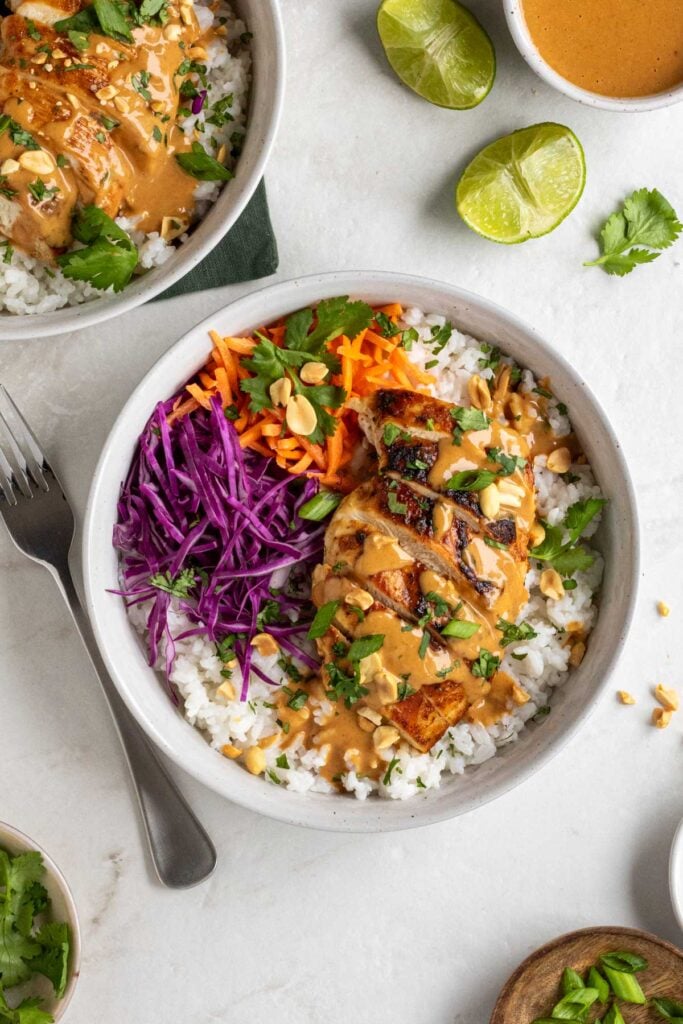 Two peanut chicken bowls with with a fork, a jar of Thai peanut sauce, chopped peanuts, and cilantro on the side on a white background.