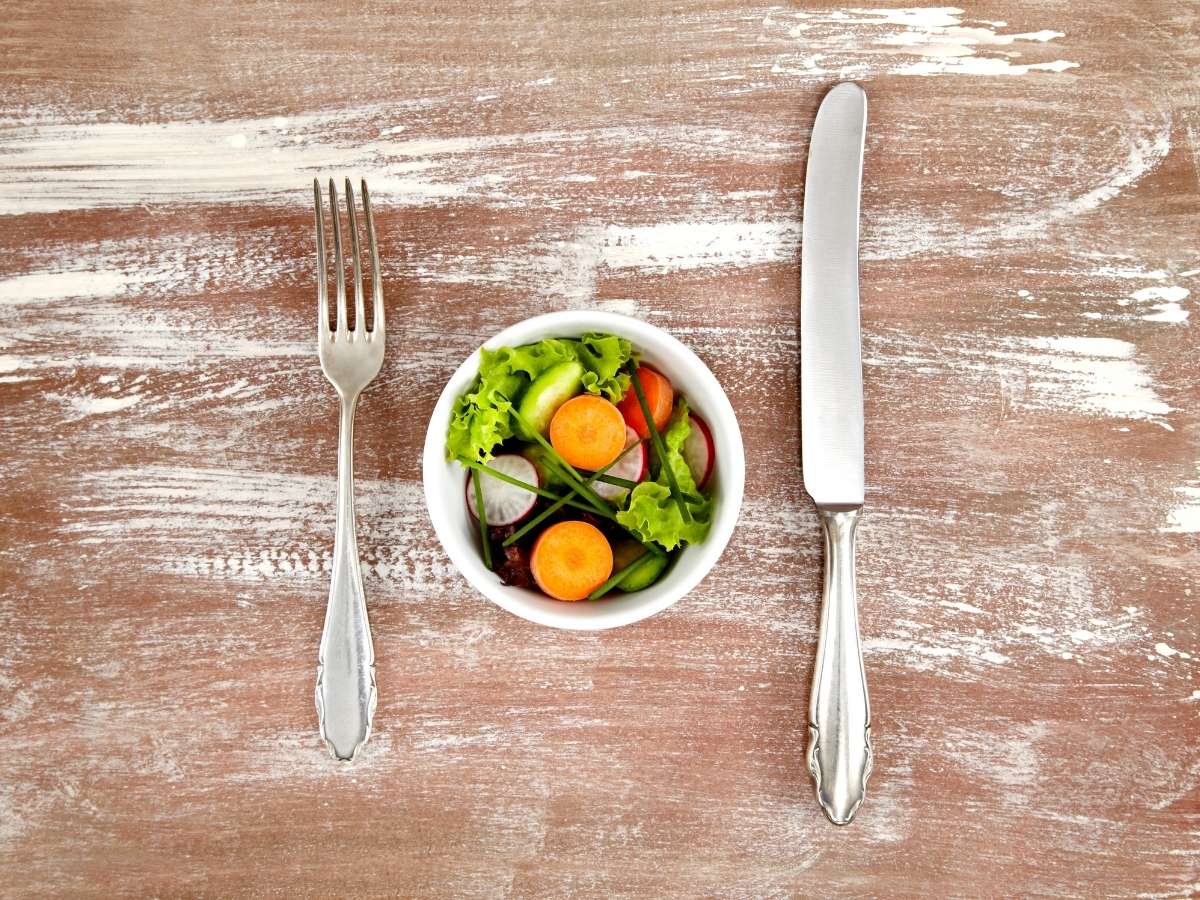Tiny white bowl of garden salad with a silver fork and knife on the side on a rustic wood background.