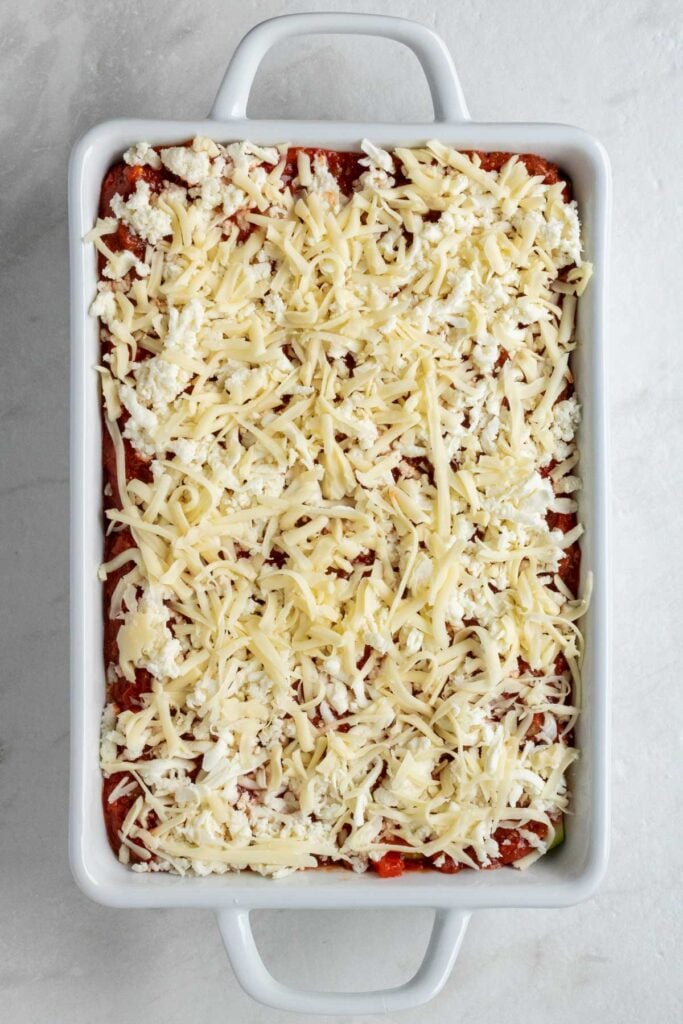 White baking dish with zucchini slices, meat sauce, spinach ricotta mixuture, and more zucchini slices covered in shredded mozzarella cheese.