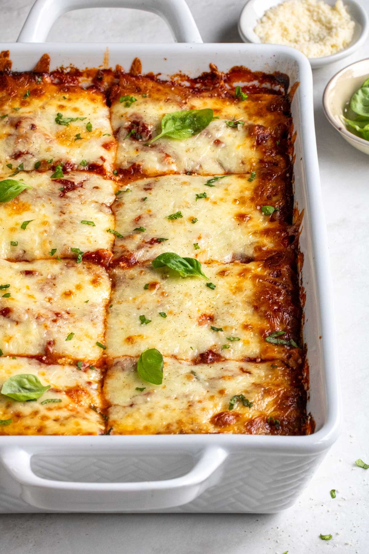 White baking dish of zucchini lasagna with a bowl of fresh basil and parmesan in the background on a white background..