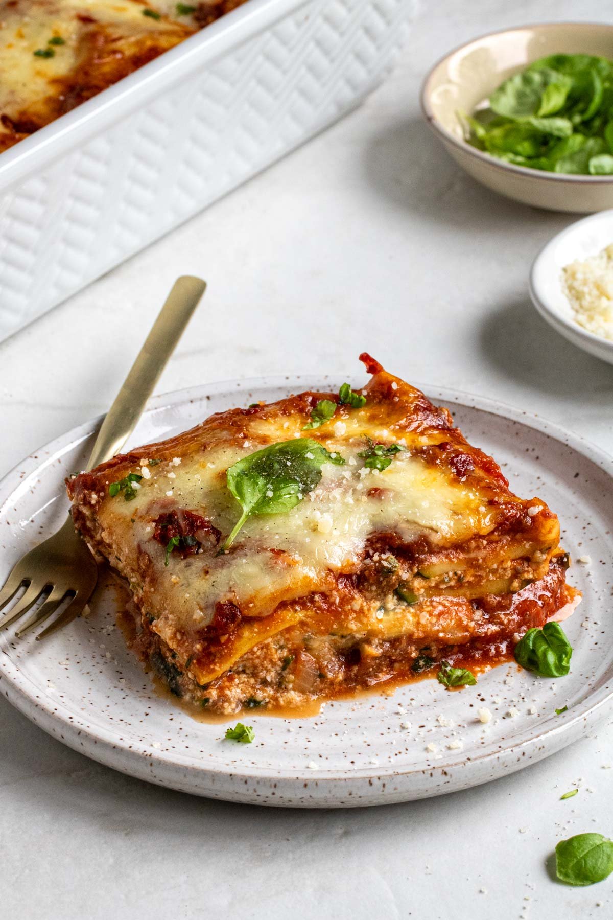Close up of a slice of zucchini lasagna on a beige plate with grated parmesan and fresh basil on top with a gold fork on a white background.