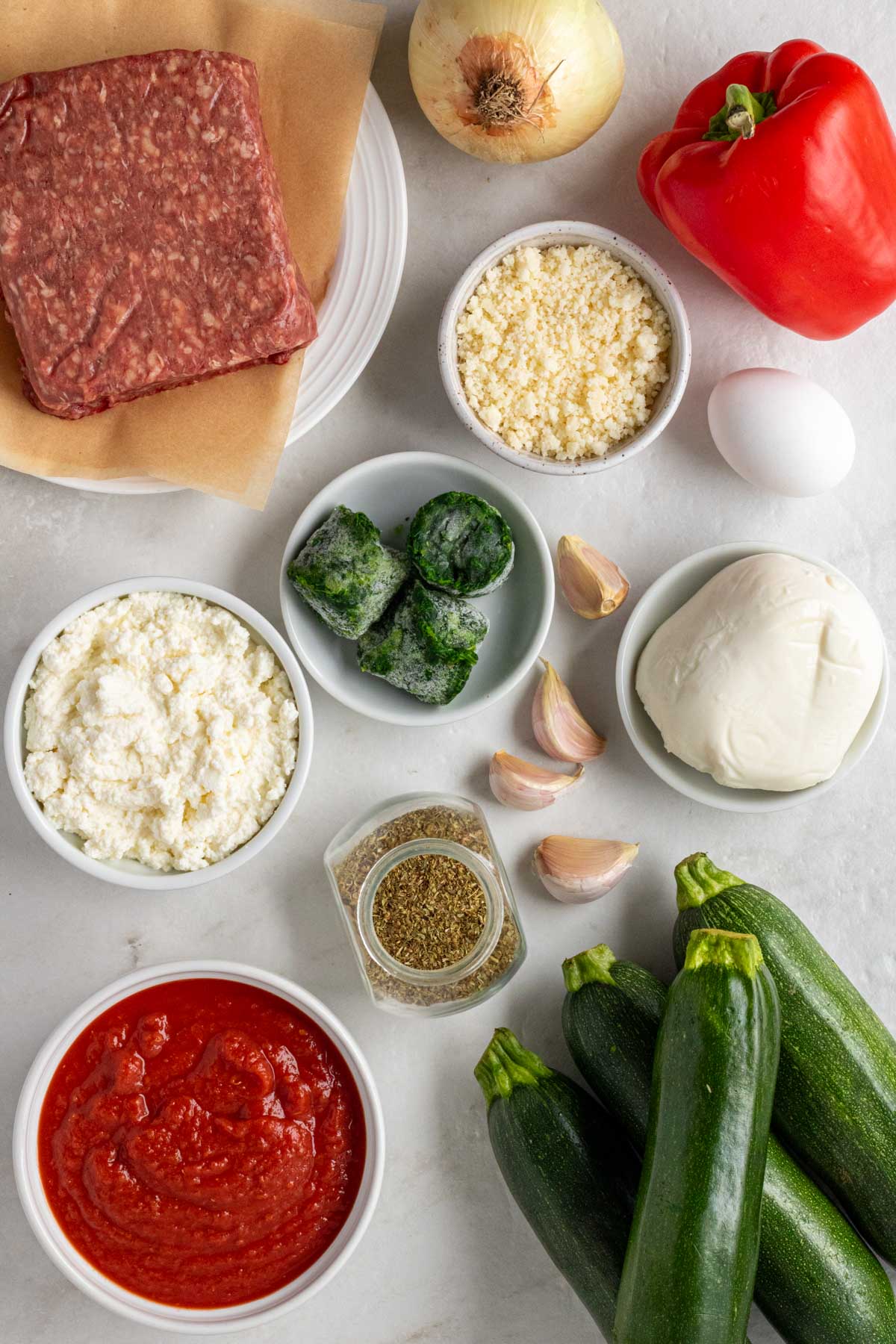 Plate of ground beef, onion, red bell pepper, bowl of freshly grated parmesan, egg, garlic cloves, bowl of ricotta, bowl of mozzarella, jar of dried oregano, bowl of crushed tomatoes, and fresh zucchini on a white background.