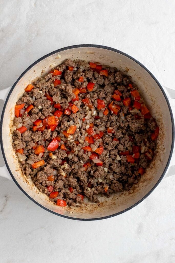Large pot with cooked ground beef, onion, red bell peppers, garlic, dried oregano, salt, and pepper on a white background.