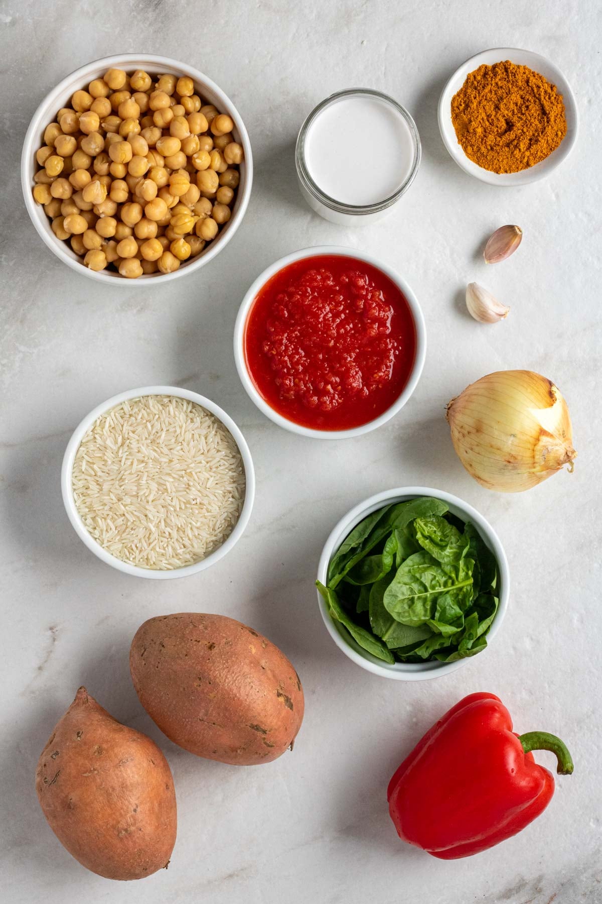Bowl of chickpeas, coconut milk, diced tomatoes, white rice, and spinach, small bowl curry powder, crushed red pepper flakes, and salt, with sweet potatoes, bell pepper, onion, garlic, and cilantro on the side.