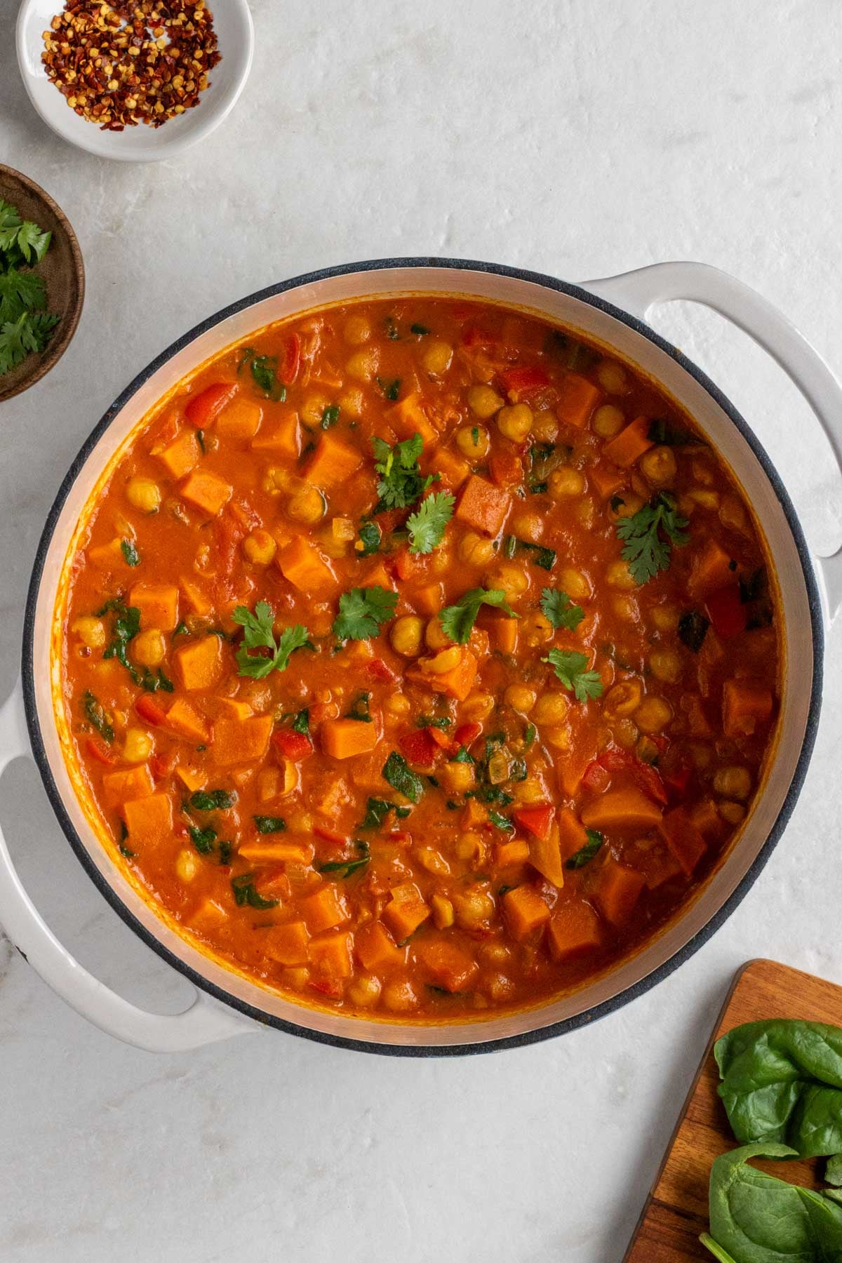 Large bot of chickpea curry with fresh spinach on a cutting board and a small white bowl of crushed red pepper flakes and a small wood bowl of cilantro on the side on a white background.