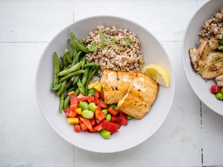 White bowl with grilled salmon, buckwheat groats, green beans, and edamame and bell pepper salad on a white background.