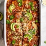 White baking dish filled with chicken enchilada casserole with avocado slices, jalapeño slices, and cilantro on top with a tea towel, a spatula, and bowls of salsa, sour cream, jalapeño, cotija cheese, and cilantro on the side on a white background.