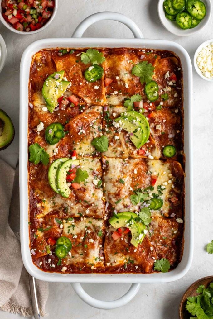 White baking dish filled with chicken enchilada casserole with avocado slices, jalapeño slices, and cilantro on top with a tea towel, a spatula, and bowls of salsa, sour cream, jalapeño, cotija cheese, and cilantro on the side on a white background.