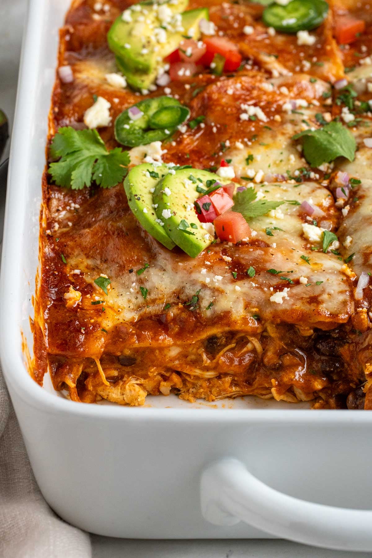 Close up of a baking dish of chicken enchilada casserole with a slice missing with avocado and jalapeño slices on top.