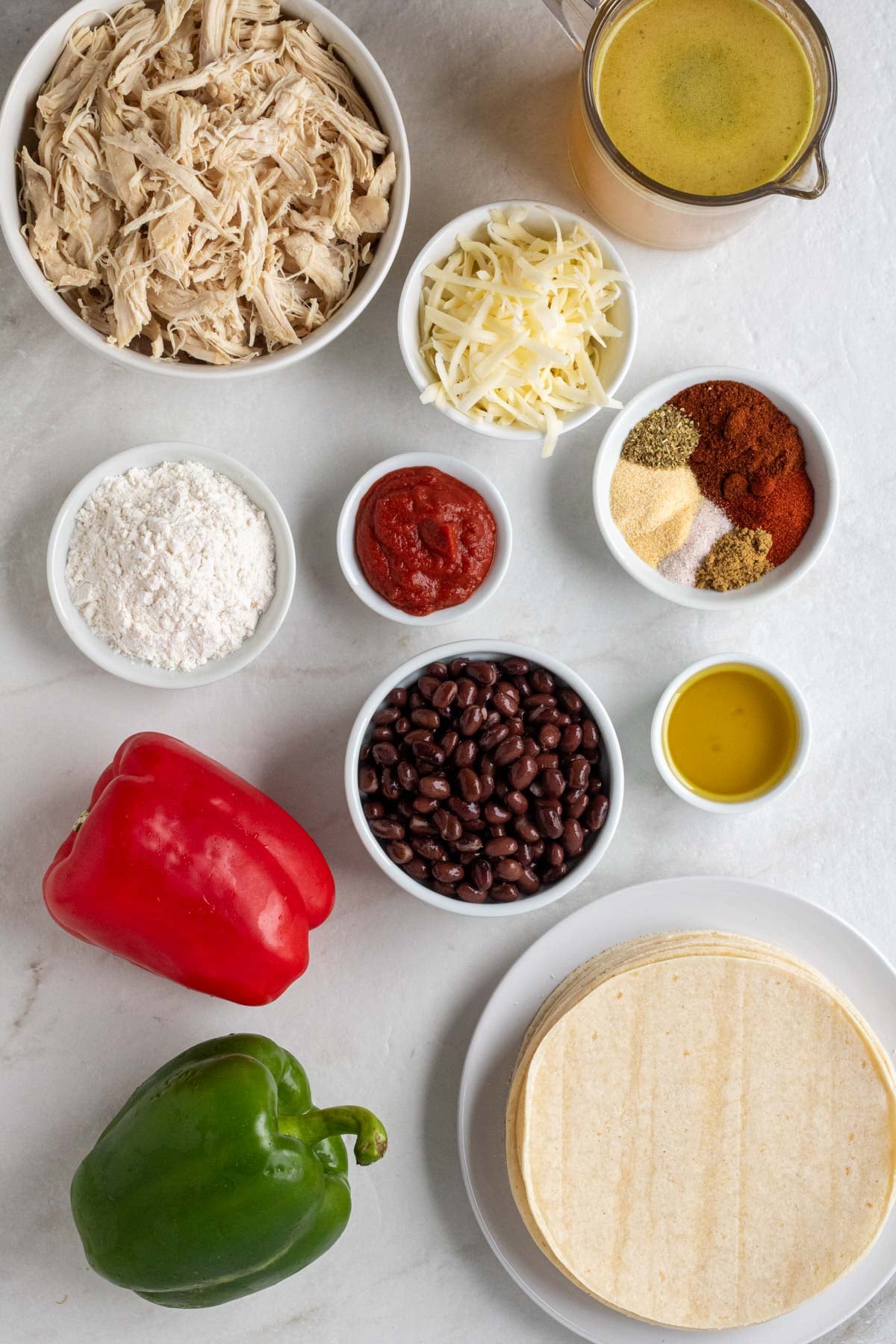 Bowls of shredded chicken, shredded cheese, flour, tomato paste, spices, black beans, and olive oil, with a container of chicken broth, bell peppers and a plate of corn tortillas on a white background.
