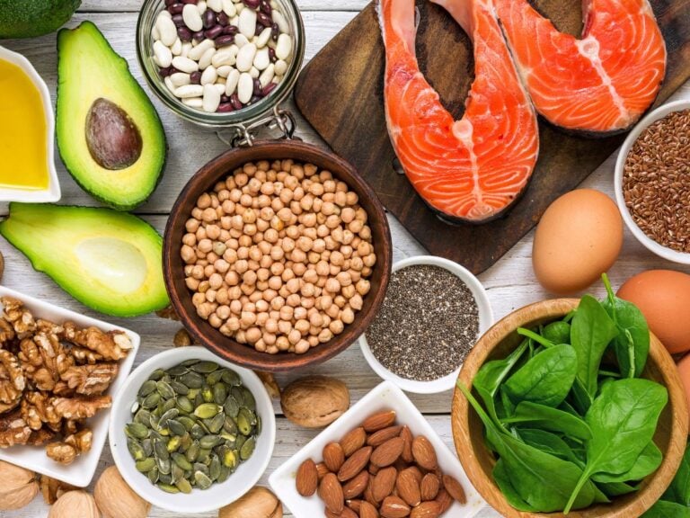 Canola oil, avocados, walnuts, beans, soybeans, pumpkin seeds, salmon fillet, chia seeds, eggs, flax seeds, and spinach on a white wood background.