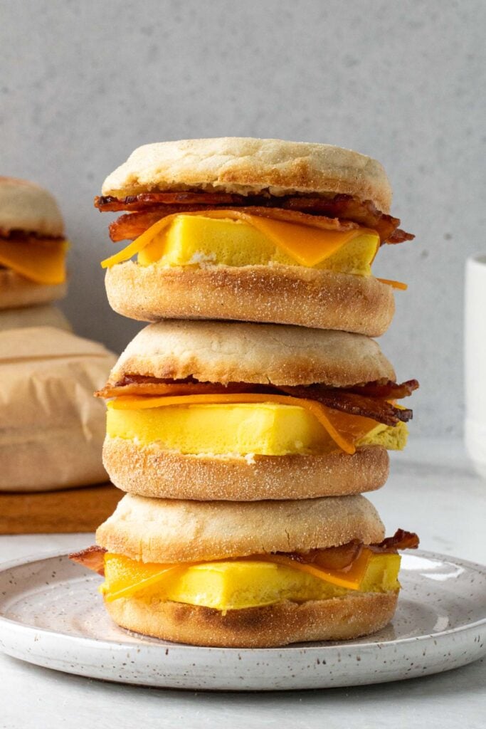Three meal prep breakfast sandwiches stacked on a plate with a breakfast egg sandwich wrapped in parchment paper in the background.