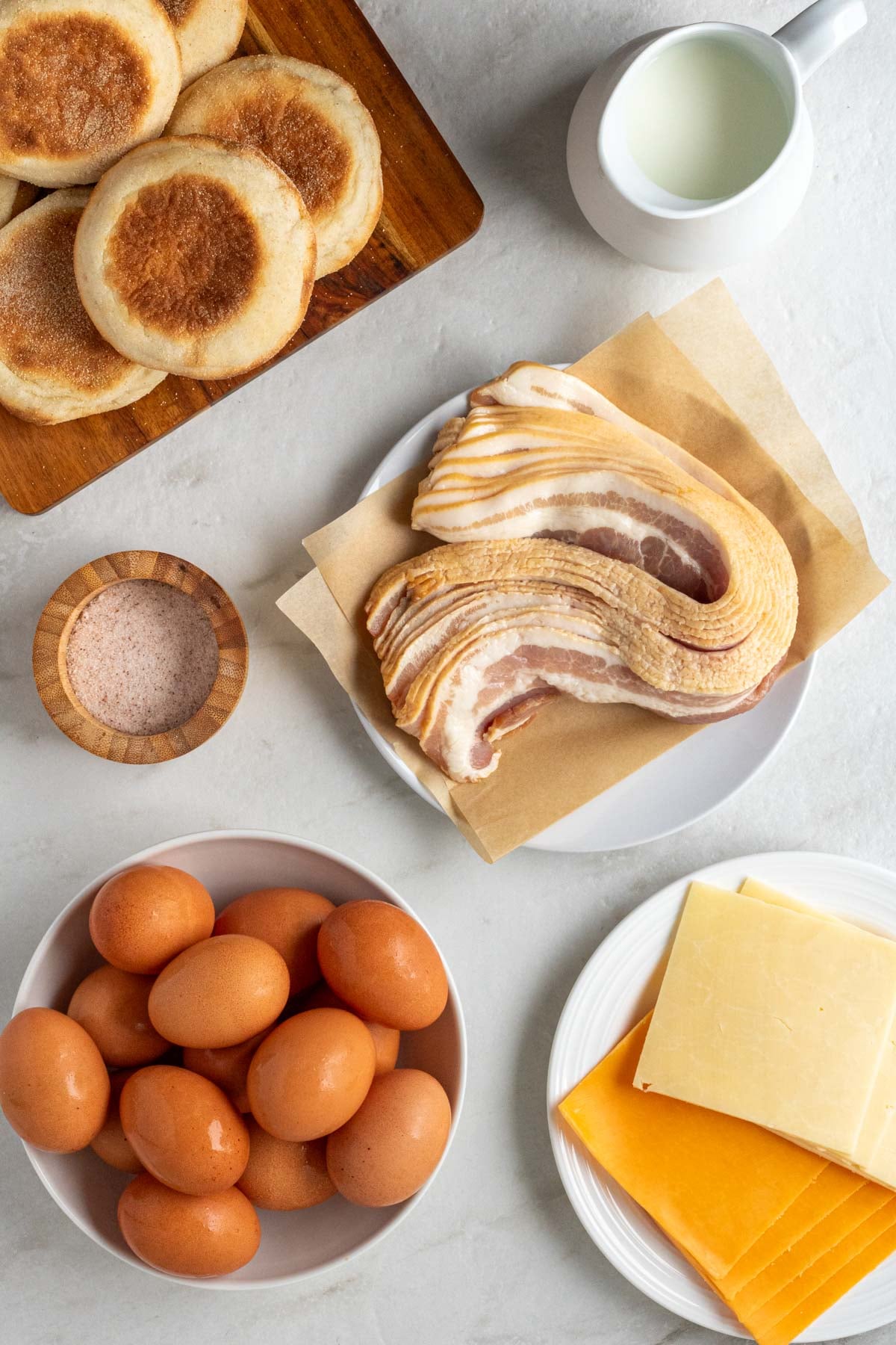 Bowl of eggs, plate of bacon, plate of cheese slices, jar of milk, and bowl of salt on a white background.