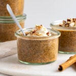 Jar of pumpkin spice chia pudding with a dollop of yogurt and chopped pecans on top with cinnamon sticks on the side on a white background.
