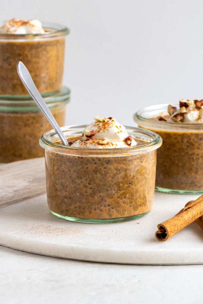 Jar of pumpkin spice chia pudding with a dollop of yogurt and chopped pecans on top with cinnamon sticks on the side on a white background.