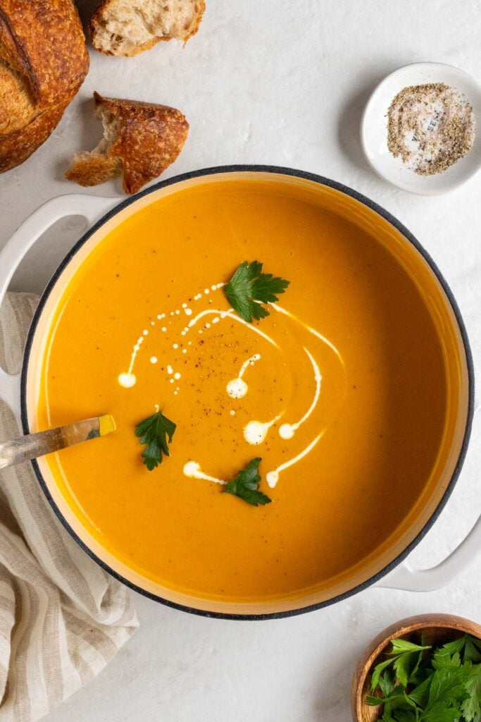 Large pot of roasted butternut squash and sweet potato soup with heavy cream and fresh parsley on top with a silver serving spoon and a loaf of sourdough bread, a bowl of salt and pepper, and beige tea towel on a white background.