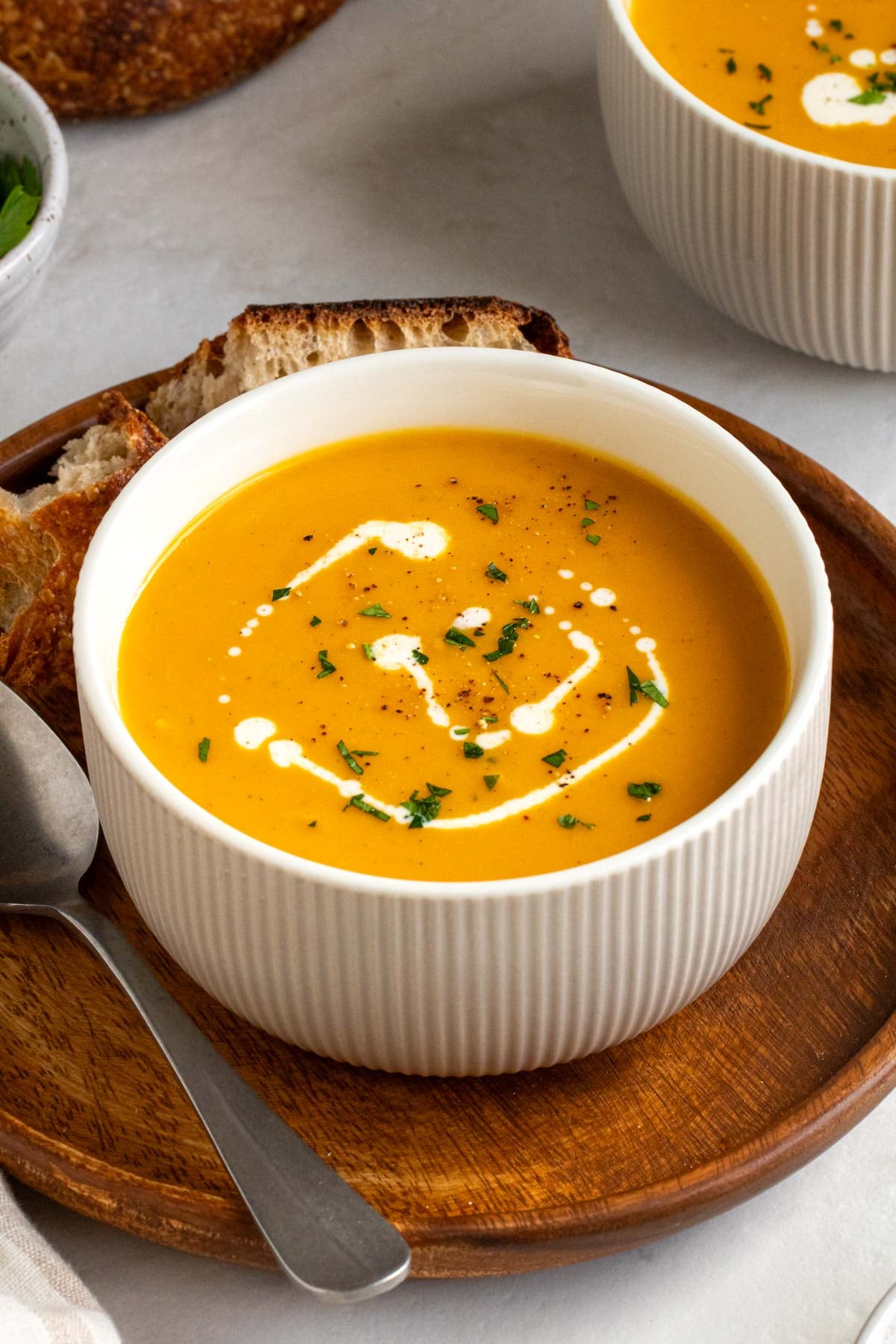 Close up of a bowl of sweet potato and butternut squash soup with cream and parsley on top on a wood plate with a silver spoon on a white background.