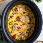 Crockpot filled with slow cooker Tuscan chicken with a bowl of pasta, chopped parsley, and shredded parmesan cheese on the side with a green tea towel on a white background.