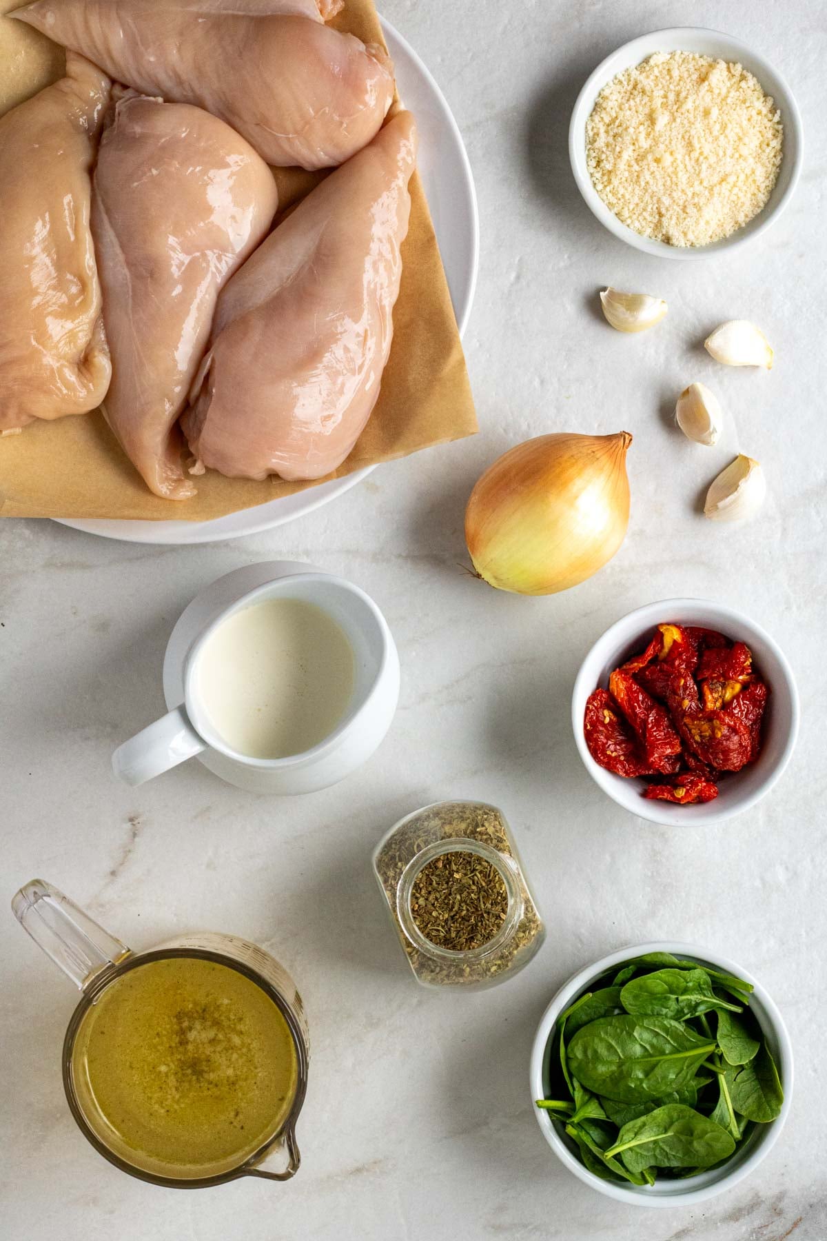 Plate of chicken breasts with a bowl of parmesan, sun-dried tomatoes, and spinach with a jar of Italian seasoning, pot of broth, pot of cream, with an onion and garlic cloves on a white background.