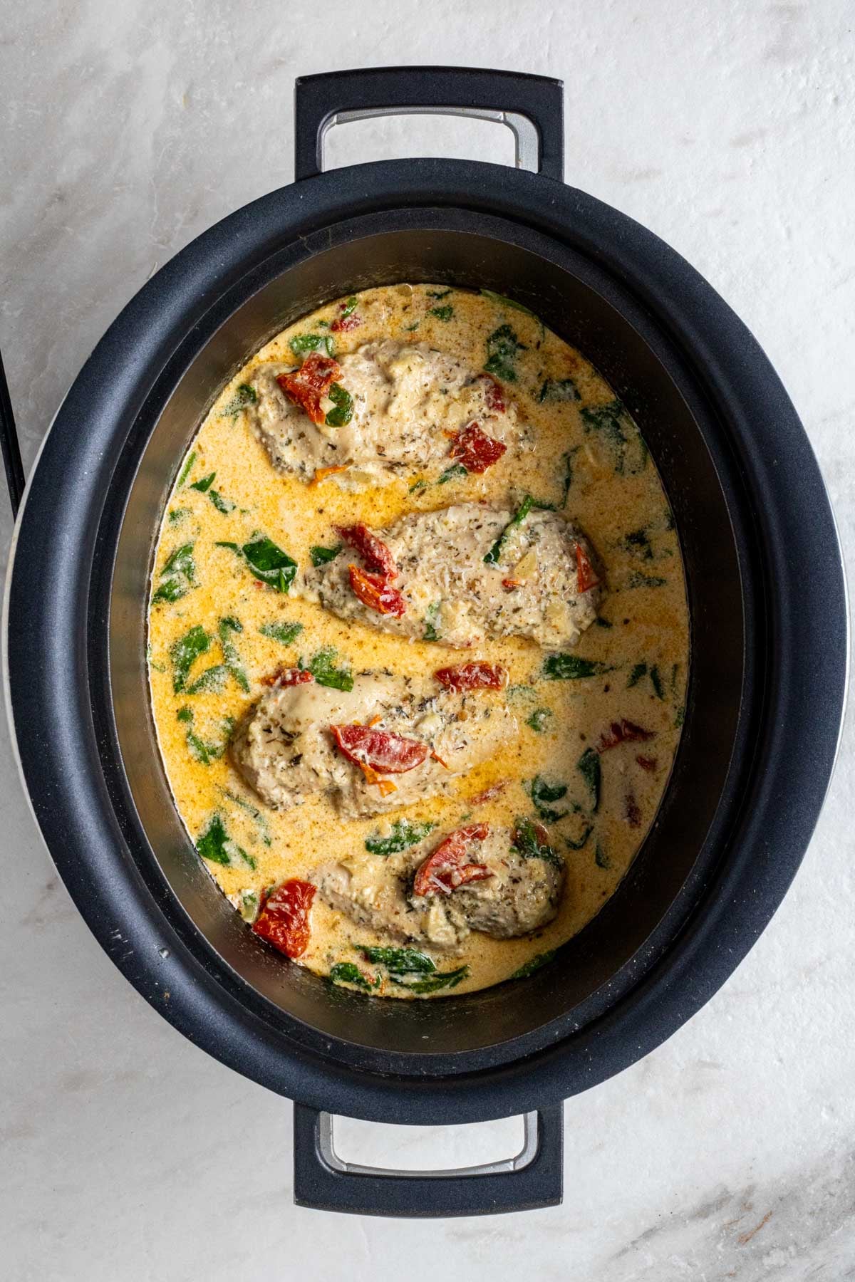 Slow cooker filled with a Tuscan chicken crockpot recipe with parmesan and parsley on top on a white background.