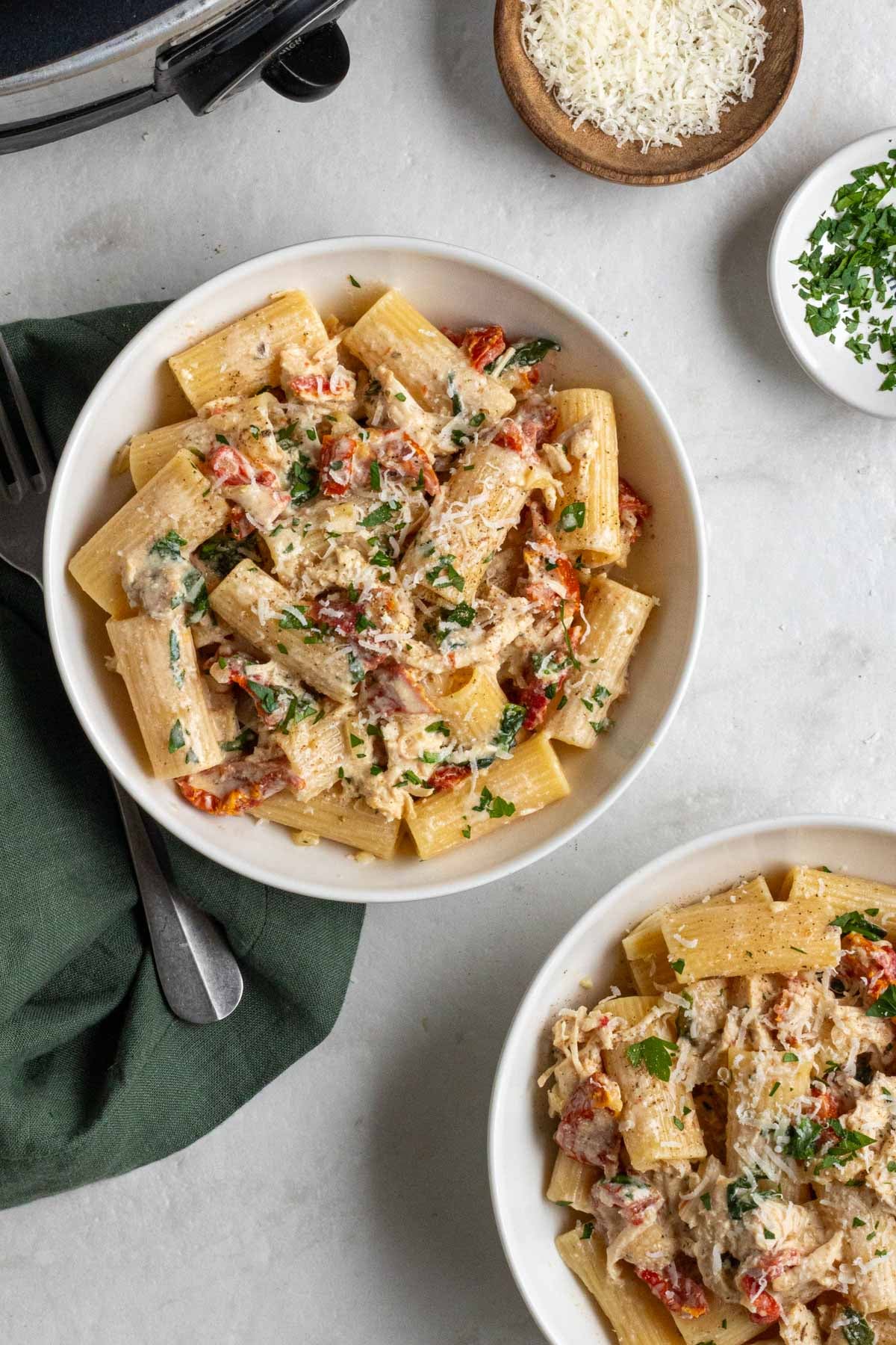 Two bowls of crockpot Tuscan chicken recipe with pasta with a silver fork and green tea towel on a white background.
