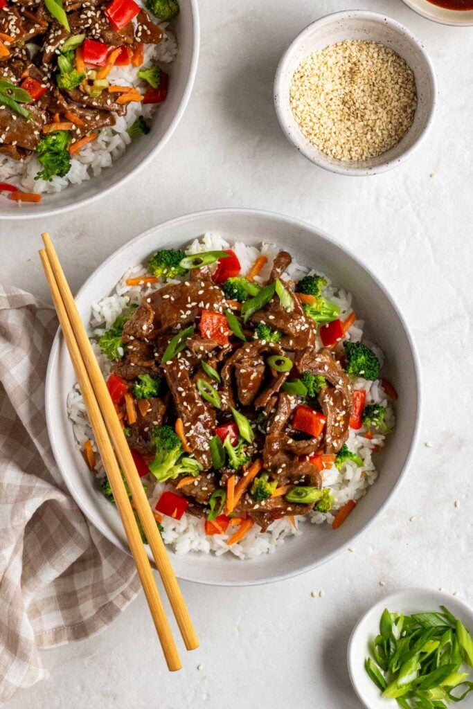 Two teriyaki beef bowls with chopsticks with a bowl of sesame seeds and sliced green onion on a white background.