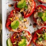 Vegetarian stuffed peppers in large baking dish with sliced avocado and fresh cilantro on top and lime wedges on the side.