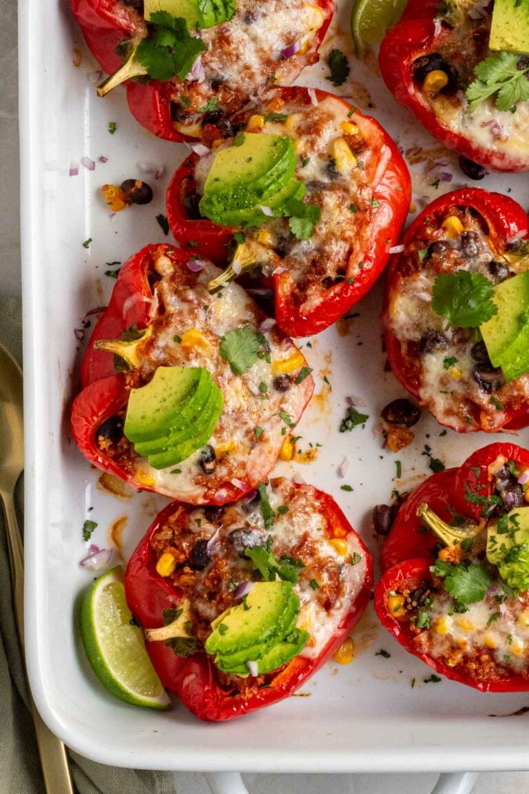 Vegetarian stuffed peppers in large baking dish with sliced avocado and fresh cilantro on top and lime wedges on the side.