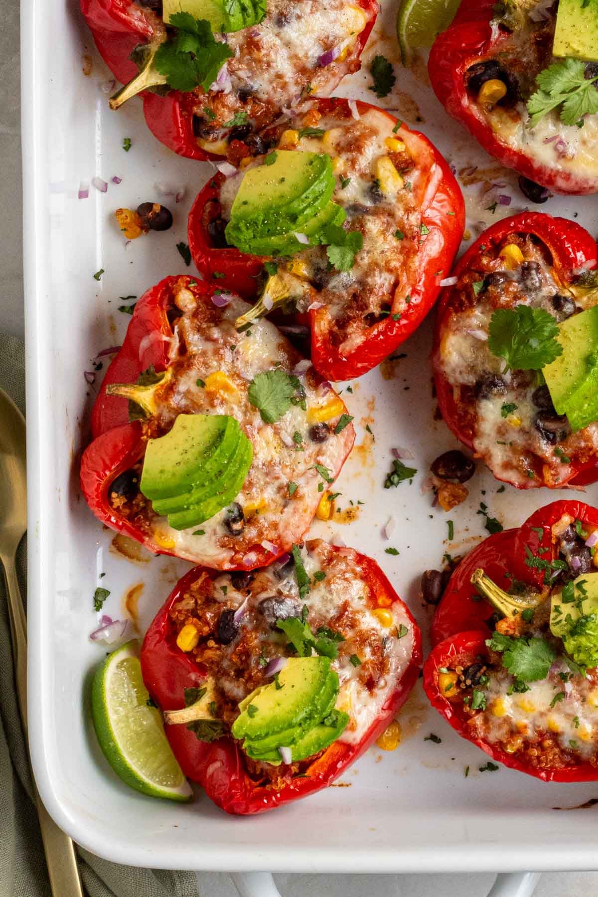 Vegetarian stuffed peppers in large baking dish with sliced avocado and fresh cilantro on top and lime wedges on the side.