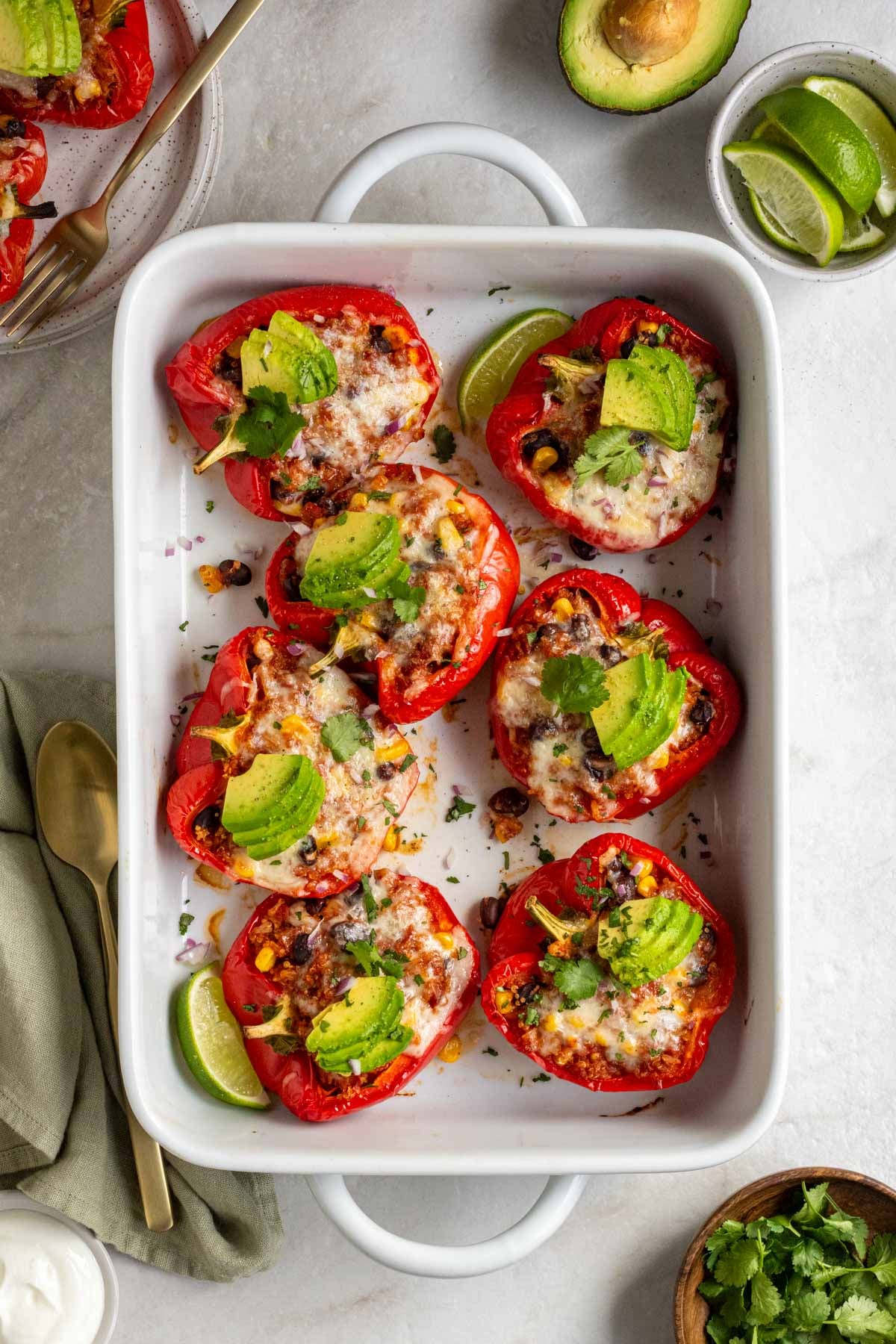 A large casserole dish filled with vegetarian stuffed bell peppers with a stack of plates, half an avocado, a bowl of lime wedges, wood bowl of fresh cilantro, with a tea towel and gold serving spoon on a white background. 
