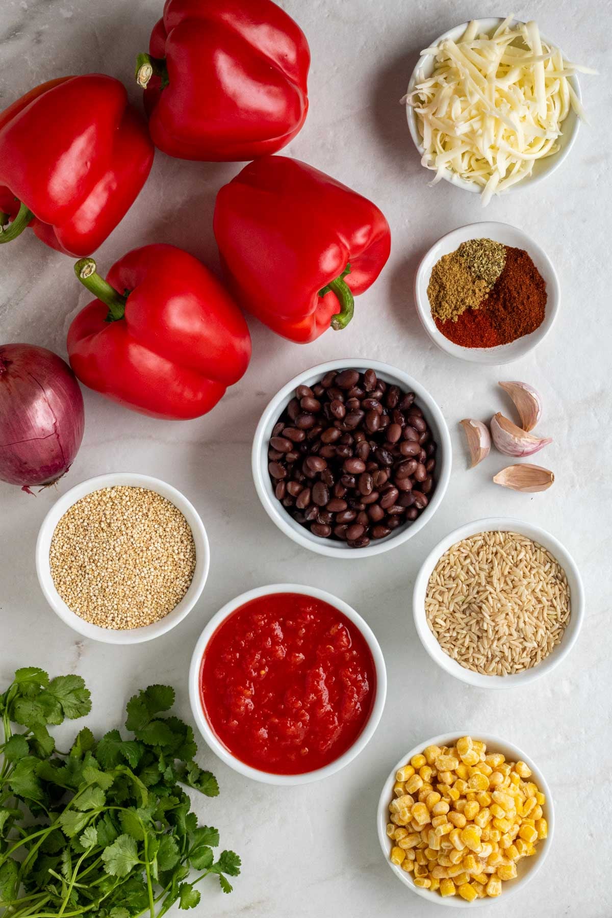 Red bell peppers, red onion, shredded cheese, spices, garlic cloves, black beans, brown rice, quinoa, diced tomatoes, frozen corn, and fresh cilantro on a white background. 