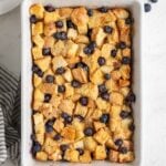 White baking dish of blueberry French toast casserole with powdered sugar sprinkled on top, fresh blueberries, fresh mint, a stripped blue tea towel and silver spatula on the side on the white background.